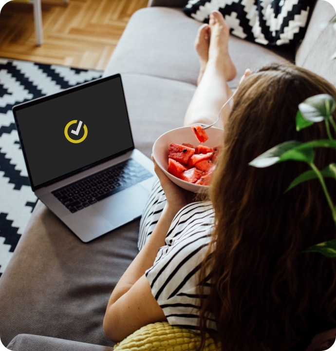 Donna sdraiata su un divano che mangia della frutta con accanto un computer portatile che mostra il logo Norton